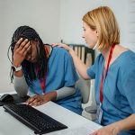 A nurse colleague places a supportive hand on the shoulder of a visibly stressed nurse at a hospital workstation