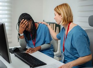 A nurse colleague places a supportive hand on the shoulder of a visibly stressed nurse at a hospital workstation