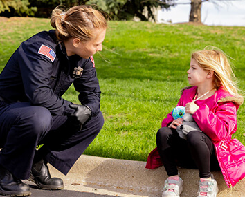 First responder in uniform comforting a young child sitting on a sidewalk.