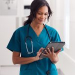 A nurse in teal scrubs walks down a hallway looking at a tablet.